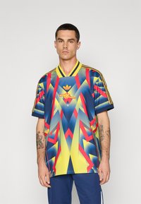 Sportovní dres s modrým podkladem a geometrickými vzory v žluté, červené a zelené barvě. Doplňuje ho žlutý véčkový výstřih a tři pruhy na rukávech.