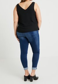Top negro sin mangas con escote en V en la parte posterior, combinado con jeans ajustados de mezclilla oscura enrollados en los puños y zapatos de tacón cuadrado negros.