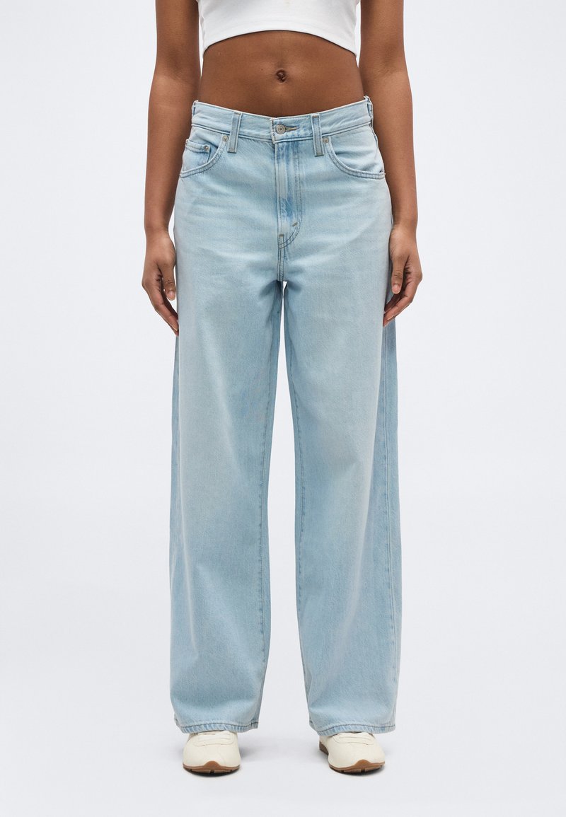 Personne portant un jean large bleu clair, un haut court blanc et des baskets blanches, debout devant un fond clair uni.