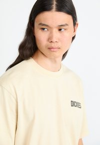 Beige bomulds t-shirt med korte ærmer, der har et lille sort "DICKIES" logo på venstre bryst. Glat tekstur, enkelt design.