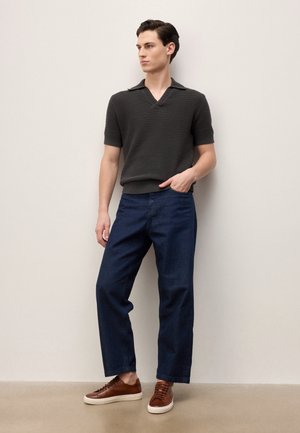 Junger Mann, der ein dunkelgraues, kurzärmeliges Strickshirt, dunkelblaue Jeans und braune Turnschuhe trägt und vor einer hellen Wand steht.