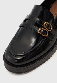 Svart lakkert skinn loafer med doble spenneaksenter, avrundet tå og teksturert gummisåle med vevd detaljering rundt kanten.
