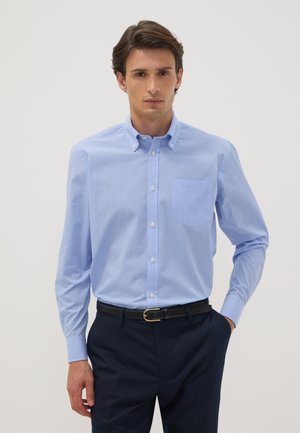 Mann trägt ein hellblau kariertes Hemd, das in dunkle marineblaue Hose mit schwarzem Gürtel gesteckt ist, und steht vor einem einfarbigen Hintergrund.