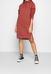 Mikina s kapucí v rezavé barvě, vyrobená z měkkého materiálu. Má dlouhé rukávy, boční rozparky a je nošena s černými vysokými teniskami.