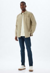 Camisa de botones marrón claro, camiseta blanca debajo, jeans azules y zapatillas crema con suelas marrones. Diseño minimalista y estilo casual.