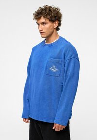 Blauwe sweatshirt met lange mouwen, ronde hals en een voorzak. De stof heeft een zachte textuur en de zak heeft geborduurde details.