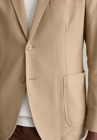Blazer beige in tessuto testurizzato, dotato di chiusure con due bottoni, una tasca sul petto e tasche laterali, che mostra una vestibilità sartoriale e linee pulite.