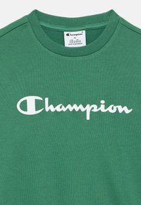 Πράσινο βαμβακερό t-shirt με στρογγυλή λαιμόκοψη. Διαθέτει το χαρακτηριστικό λευκό λογότυπο "Champion" στο μπροστινό μέρος. Μαλακό, υφασμάτινο ύφασμα με υφή.