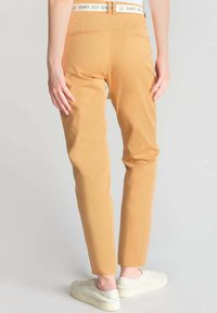 Chinos giallo senape slim-fit con tasche posteriori e cintura bianca, abbinati a sneakers bianche per un look casual.