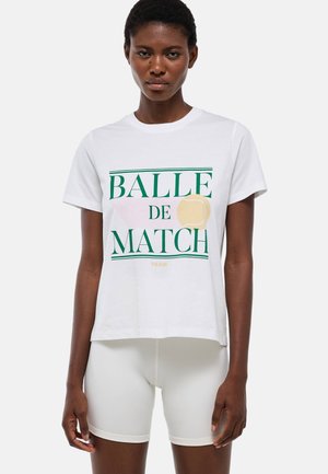 Femme portant un t-shirt blanc avec un texte vert "Balle de Match" et un graphisme de balle de tennis, associé à un short de cyclisme blanc.