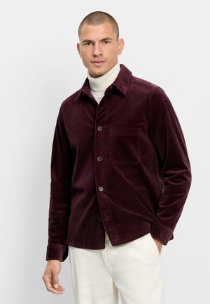 Man wearing a dark red corduroy button-up shirt over a white turtleneck and white pants, standing against a plain light background.