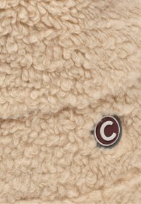 Textil bej pufos cu o suprafață texturată, în bucle. Prezintă un logo circular cu un fundal vișiniu și litera albă "C" atașată.
