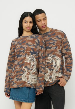 Młody mężczyzna i kobieta w pasujących brązowych swetrach moro z dużymi białymi grafikami kobry, stojący blisko obok siebie.