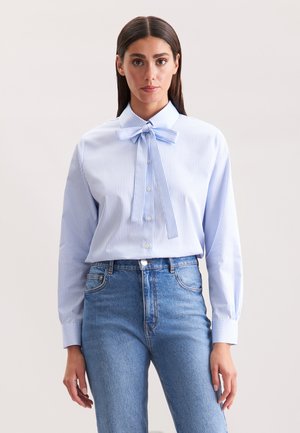 Blusa azzurra a strisce con collo a fiocco, maniche lunghe e chiusura a bottoni. Indossata con jeans in denim blu a vita alta.