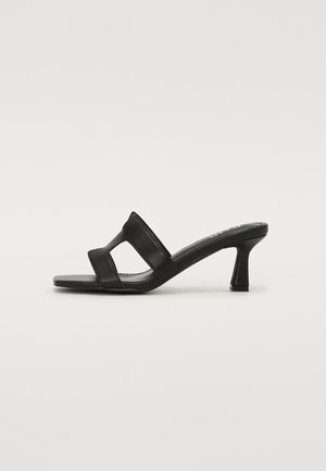 Mules noirs à talons en cuir lisse avec deux larges brides et un talon cylindrique. La semelle est plate et texturée pour une meilleure adhérence.