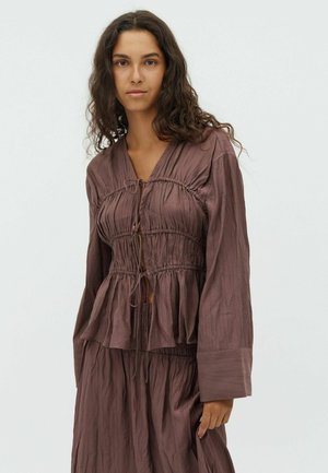 Brown, lightweight blouse with a textured surface. Features a V-neck, gathered waist, and long sleeves. Ties at the front.