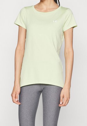 Kvinde iført en lysegrøn kortærmet sportstrøje og grå tætsiddende leggings, stående mod en ensfarvet lys baggrund.