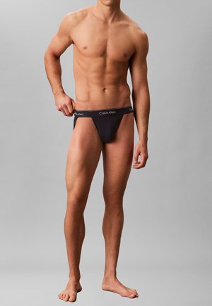 Athletisches männliches Model trägt schwarze Calvin Klein Jockstrap-Unterwäsche, justiert den Bund und steht barfuß vor einem schlichten grauen Hintergrund.