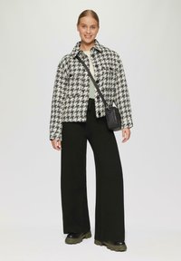Zwart-witte houndstooth jas met een kraag, gedragen over een licht topje en brede zwarte broek. Details zijn een kleine zwarte crossbody tas en chunky schoenen.