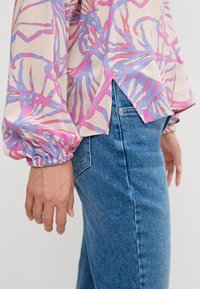 Blouse légère en tissu doux, présentant des motifs abstraits roses et bleus, manches longues bouffantes avec poignets élastiques, associée à un jean en denim bleu.