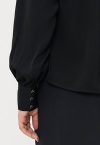 Blusa nera con maniche lunghe a sbuffo, orlo arrotondato e polsini con bottoni, caratterizzati da quattro piccoli bottoni circolari. Tessuto liscio e leggero.