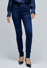 Jeans skinny de mezclilla azul oscuro con una textura suave, diseño de cinco bolsillos y cintura alta. Combinados con tacones negros de punta afilada.