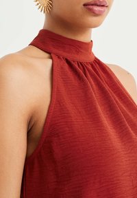 Rustfärgad halterneck-topp i texturerad tyg, med hög krage och flödande design, öppen axelutskärning och samlade detaljer.