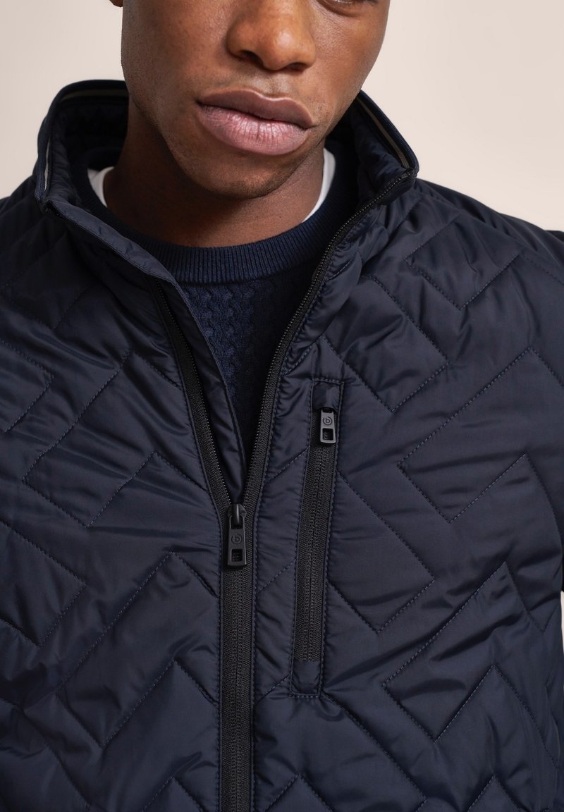 Marineblaue gesteppte Jacke mit hohem Kragen, ausgestattet mit zwei Frontreißverschlüssen und einem Rautenmuster. Das Material wirkt glatt und leicht.