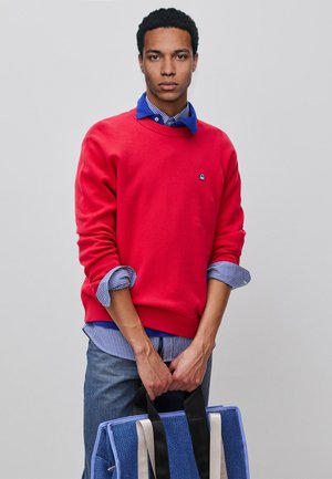 Młody mężczyzna w czerwonym swetrze na niebieskiej koszuli w kratę, trzymający niebieską i czarną torbę na ramię, stojący na jednolitym szarym tle.