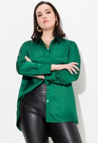 Zielona satynowa bluzka z guzikiem z przodu, długimi rękawami i luźnym krojem, zestawiona z eleganckimi czarnymi spodniami z ekoskóry.