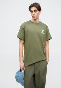 Ελαιόχρωμο βαμβακερό μπλουζάκι με κοντά μανίκια και στρογγυλή λαιμόκοψη, με μικρό λευκό λογότυπο Nike και κείμενο στο αριστερό στήθος.