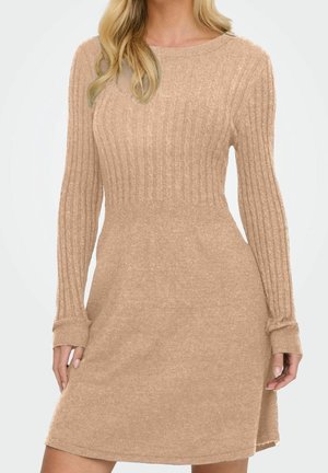 Kvinde iført en beige ribstrikket langærmet kjole med tætsiddende talje og rund hals, stående mod en hvid baggrund.
