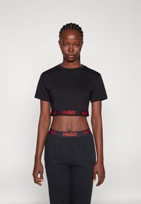 T-shirt noir court avec logo "HUGO" rouge sur l'ourlet, associé à un pantalon noir avec ceinture assortie. Matériau en coton lisse.