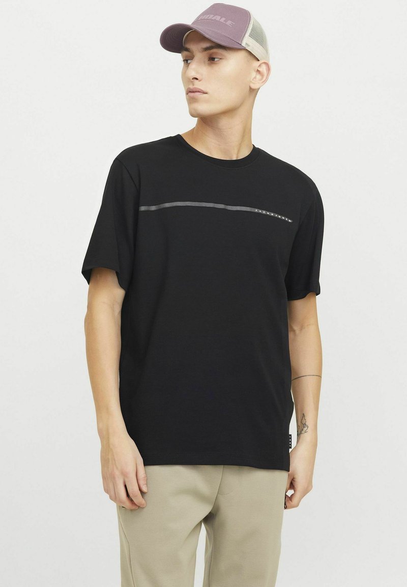 Sort kortærmet T-shirt med en horisontal grå linje og små nitter på tværs af brystet. Bæres med lysebeige bukser og en lilla kasket.