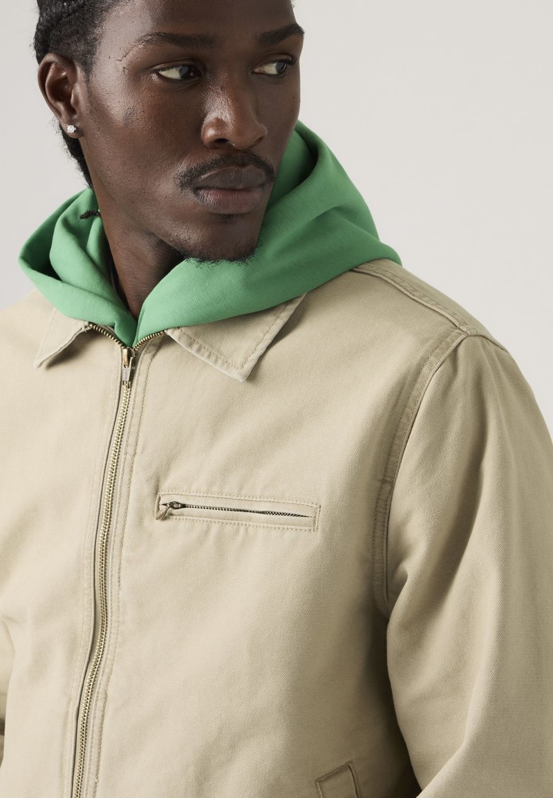 Mann trägt eine beige Jacke mit Reißverschluss über einem grünen Kapuzenpullover und schaut seitlich vor einem schlichten hellen Hintergrund.