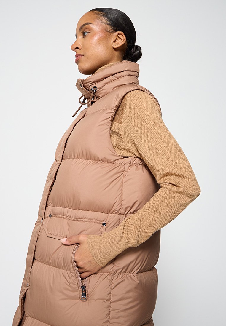 Braune Pufferweste mit hohem Kragen, Seitentaschen und Kordelzug-Details. Tragen über einem strukturierten, beigen Langarmtop.