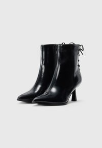 Une paire de bottines en cuir noir brillant avec des bouts pointus, une fermeture éclair sur le côté, un laçage à l'arrière et des talons fins de hauteur intermédiaire sur fond blanc.