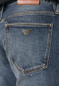Dark blue denim jeans with contrasting orange stitching, featuring a rear pocket with a gold logo accent. Brown leather waistband detail.