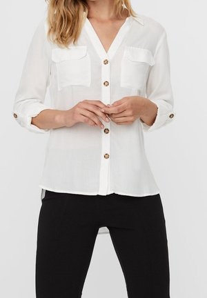 Kobieta ubrana w białą koszulę z guzikami, z dwoma kieszeniami na piersiach i podwiniętymi rękawami, w połączeniu z czarnymi spodniami.