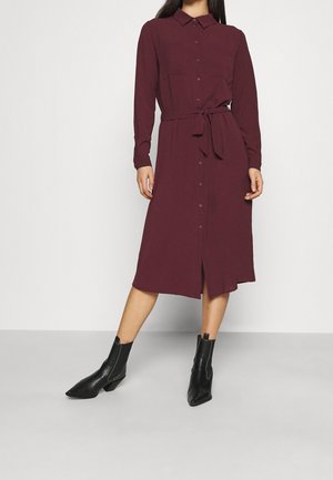 Femme portant une robe midi bordeaux à manches longues, boutonnée, avec un lien à la taille et des bottines noires, debout devant un fond clair uni.