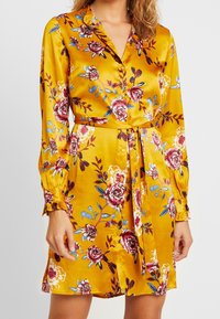Satin skjortklänning i senapsgul med blommönster och röda, blåa och vita accenter, långa ärmar och midjebälte.
