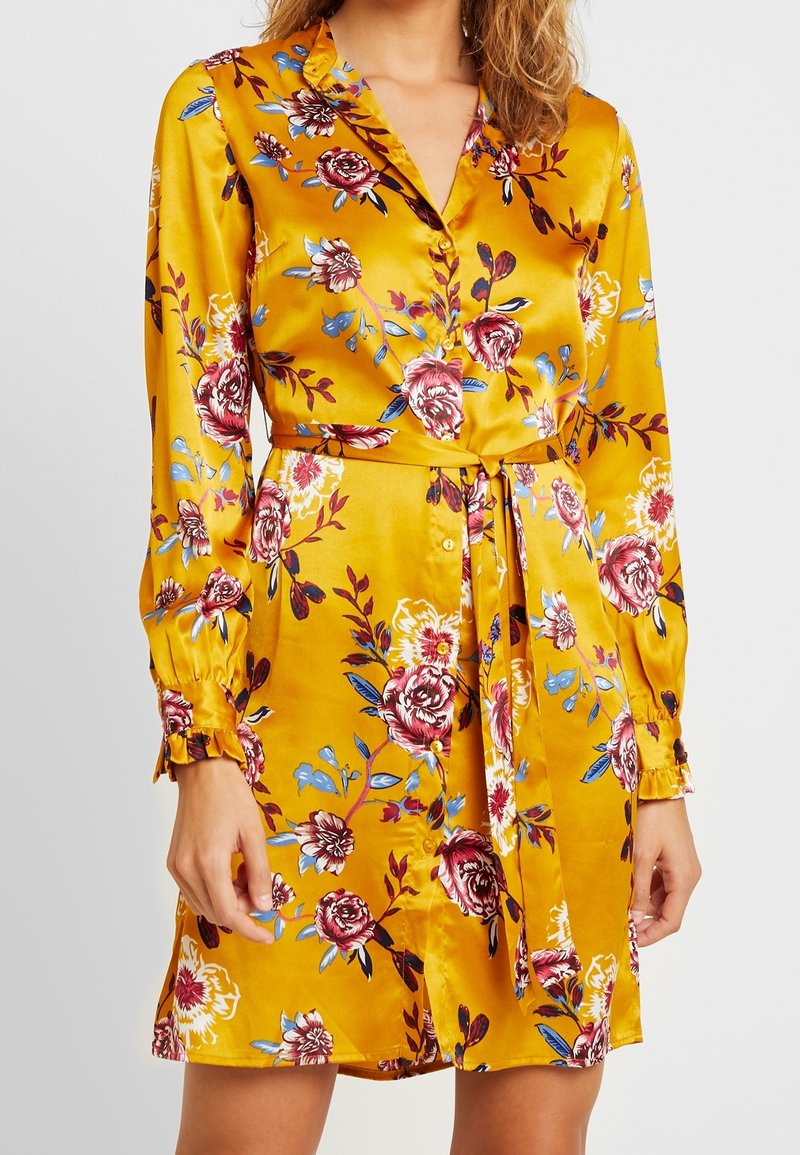 Satin skjortklänning i senapsgul med blommönster och röda, blåa och vita accenter, långa ärmar och midjebälte.