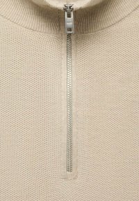 Suéter beige de cremallera con un patrón de punto texturizado, cuello alto y una cremallera plateada en el frente. El material parece suave y cálido.