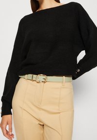 Pull en maille noire texturée avec un design décolleté sur les épaules, associé à un pantalon beige taille haute et une ceinture vert clair avec une boucle dorée.