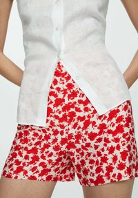 Bílá lněná košile na knoflíky spárovaná s červenými kratšími kalhotami s květinovým vzorem na světlém pozadí. Hlavními rysy jsou květinový design a lehká textura.