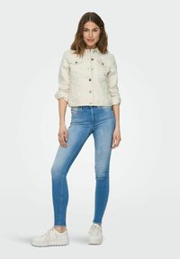 Light beige denim jacket featuring a fitted design, button closure, and two front pockets, paired with slim-fit blue jeans and white sneakers.