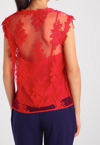 Rød gennemsigtig bluse med detaljer i blomsterlacet, bølget halsudskæring og cap sleeves; lagdelt design over en solid bund.