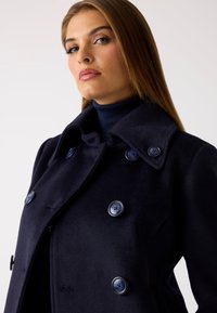 Abrigo de marina de doble botonadura con grandes botones azules y un amplio cuello, hecho de una tela suave, que presenta una silueta ajustada y un cuello alto acanalado debajo.