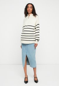 Femme avec des cheveux bouclés portant un pull blanc à zip demi-ouvert avec des rayures noires, une jupe midi en denim bleu clair avec une fente à l'avant, et des talons noirs.