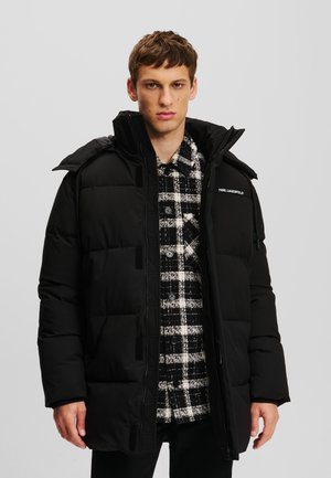 Black puffer jacket with a high collar and visible zip, layered over a checkered black and white shirt. Features sleek, textured fabric.